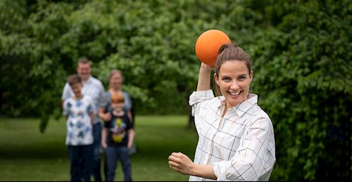 Sofie Østergaard står i en have med en bold i hånden - klar til at kaste. I baggrunden står en familie på fire klar til at lege i haven.